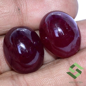 May include: Two oval-shaped, deep red gemstones with a high gloss finish. The gemstones are set against a neutral background.