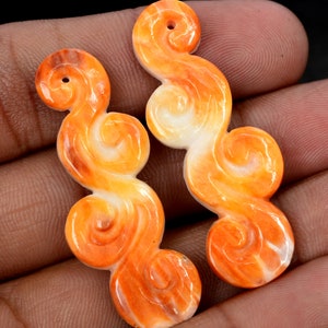 May include: Two orange and white carved shell pendants in the shape of swirling clouds. The pendants have a hole at the top for stringing.