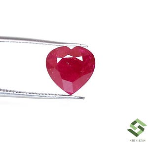 Peut inclure: Un rubis en forme de cœur, facetté, tenu par une pince. Le rubis est d'un rouge profond et présente une petite coupe triangulaire en haut. La pierre précieuse est posée sur un fond blanc.