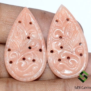 May include: Two peach-colored, teardrop-shaped gemstone carvings. Each carving features a detailed floral and leaf design with several small, evenly spaced holes. The carvings are displayed against a neutral background.