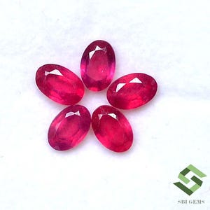 May include: Five oval-cut, vibrant red rubies arranged in a starburst pattern. Each gemstone displays facets that catch the light, creating a sparkling effect. The rubies are set against a white background, highlighting their rich color and clarity.