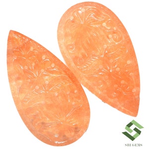 May include: Two peach-colored, teardrop-shaped gemstones with intricate floral carvings. The gemstones have a polished surface and are set against a white background. The carvings feature detailed leaves and dotted accents, showcasing the craftsmanship of the jewelry.