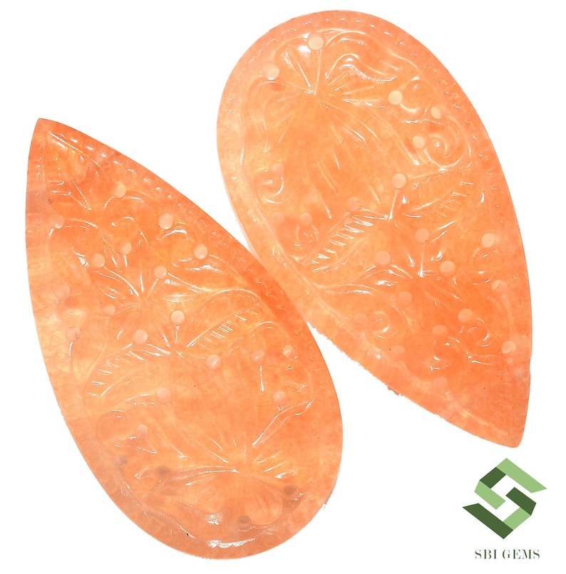 May include: Two peach-colored, teardrop-shaped gemstones with intricate floral carvings. The gemstones have a polished surface and are set against a white background. The carvings feature detailed leaves and dotted accents, showcasing the craftsmanship of the jewelry.