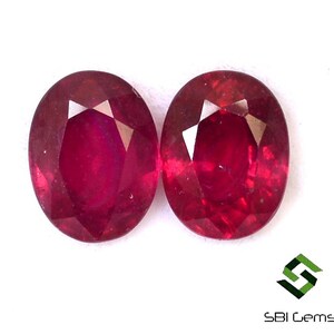 Puede incluir: Dos rubíes de corte ovalado, de color rojo intenso, sobre un fondo blanco. Las piedras preciosas tienen una superficie facetada que refleja la luz. La imagen destaca la claridad y el color de los rubíes, ideales para joyería.