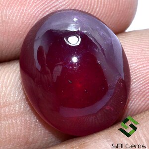 Puede incluir: Una piedra preciosa ovalada cabujón de color rojo intenso con un alto pulido. La piedra preciosa tiene un ligero tono púrpura alrededor de los bordes.