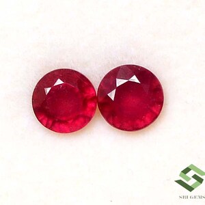 Pode incluir: Duas pedras preciosas redondas, facetadas, de cor vermelha profunda. Cada gema tem um corte brilhante com facetas triangulares que captam a luz. As gemas estão sobre um fundo branco, realçando sua cor rica e clareza. As gemas são provavelmente rubis.