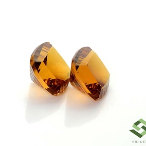 May include: Two faceted, golden-yellow gemstones are displayed against a white background. The gems have a geometric cut, reflecting light and showcasing their clarity. The gems are likely citrine or a similar gemstone.