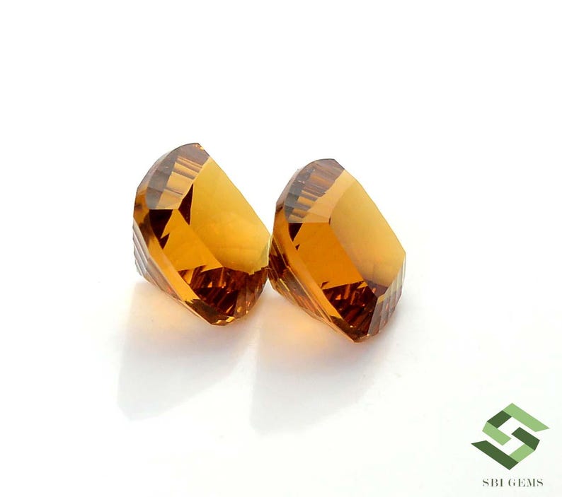 May include: Two faceted, golden-yellow gemstones are displayed against a white background. The gems have a geometric cut, reflecting light and showcasing their clarity. The gems are likely citrine or a similar gemstone.