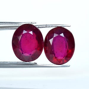 Puede incluir: Dos piedras preciosas de rubí de forma ovalada, cada una con un color rojo intenso y un corte facetado. Las piedras preciosas están sujetas por un par de pinzas.