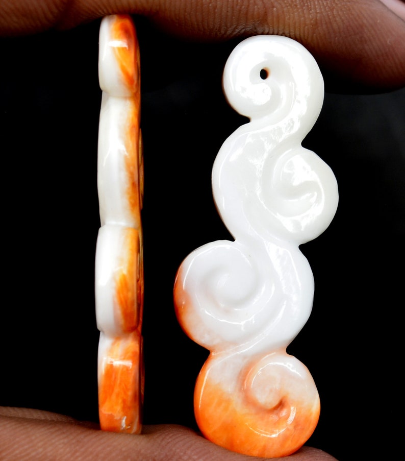 May include: A white and orange carved shell pendant in the shape of a cloud. The pendant has a hole at the top for hanging.