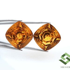 May include: Two faceted, square-cut gemstones with a warm, golden-orange hue. The gems are held by a pair of tweezers against a white background. The gems have intricate internal patterns, reflecting light. The image includes a logo in the bottom right corner.