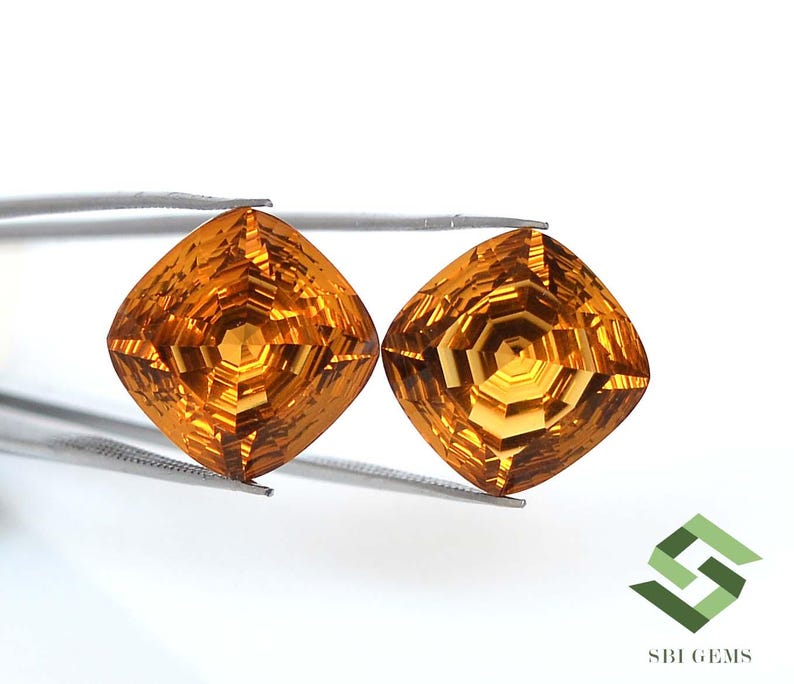 May include: Two faceted, square-cut gemstones with a warm, golden-orange hue. The gems are held by a pair of tweezers against a white background. The gems have intricate internal patterns, reflecting light. The image includes a logo in the bottom right corner.