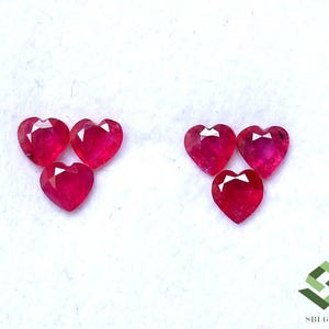 May include: Six heart-shaped, faceted rubies in a vibrant red hue are arranged in two groups of three. The gemstones are displayed against a white background, showcasing their polished surfaces and intricate cuts. The gems are likely for jewelry making.