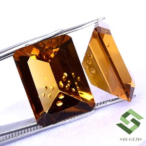 May include: Two faceted, amber-colored gemstones are held by tweezers against a white background. The larger gem is rectangular, while the smaller one is a trapezoid. The gems have internal inclusions and reflect light.