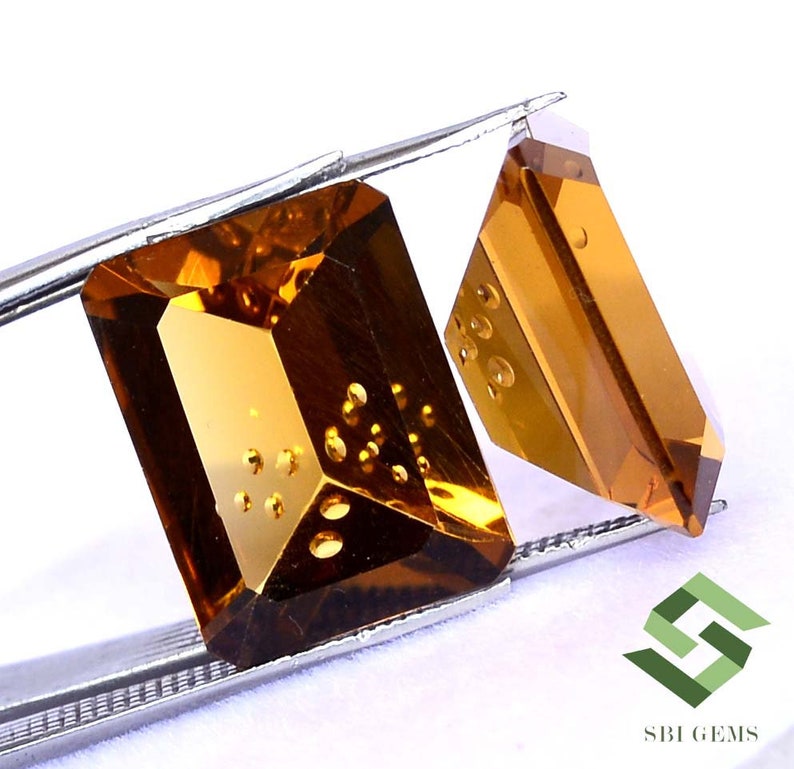 May include: Two faceted, amber-colored gemstones are held by tweezers against a white background. The larger gem is rectangular, while the smaller one is a trapezoid. The gems have internal inclusions and reflect light.