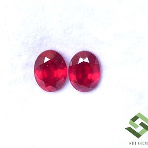 Pode incluir: Dois rubis ovais e facetados. As pedras preciosas são de cor vermelha profunda e parecem ser de alta qualidade. Os rubis são exibidos em um fundo branco, destacando sua cor e clareza. A imagem é bem iluminada, mostrando o brilho das pedras preciosas.