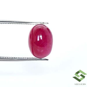 May include: A polished, oval-shaped ruby gemstone held by tweezers. The ruby is a deep, saturated red color with a smooth, reflective surface. The tweezers are silver-colored and hold the gemstone against a white background. The image is well-lit, highlighting the gem's clarity.