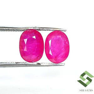 Puede incluir: Dos piedras preciosas ovaladas de color rosa, probablemente rubíes, se sostienen con unas pinzas. Las piedras preciosas son de un color rosa vibrante y profundo y tienen una superficie lisa y pulida.
