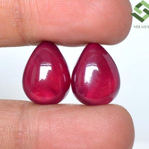 Könnte beinhalten: Zwei birnenförmige, tiefrote Rubine mit einer glatten, polierten Cabochon-Oberfläche. Die Edelsteine weisen einen satten, kräftigen Karmesinton und eine reflektierende Oberfläche auf. Das Logo von SBI GEMS ist in der oberen rechten Ecke zu sehen.