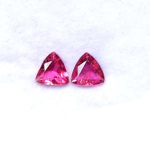 Könnte beinhalten: Zwei dreieckige, facettierte Edelsteine in leuchtendem Rosa. Die Edelsteine sind vor einem weißen Hintergrund platziert, der ihre polierten Oberflächen und scharfen Winkel hervorhebt. Diese Edelsteine sind wahrscheinlich für die Schmuckherstellung.