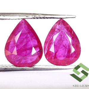 May include: Two pear-shaped ruby gemstones, each with a deep pink hue, are held by a pair of tweezers. The gemstones are faceted and have a brilliant shine.