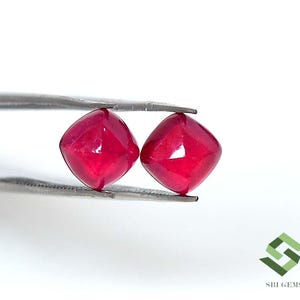 以下が含まれることがあります： 金属製のピンセットで保持された、鮮やかな深紅色のクッションカットルビー2石。宝石は滑らかで磨かれた表面を持ち、角が丸みを帯びた正方形の形状で、全体に豊かで彩度の高い深紅の色合いを呈しています。