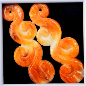 May include: Two orange and white carved jade pendants in the shape of swirling clouds. The pendants have a hole at the top for hanging.