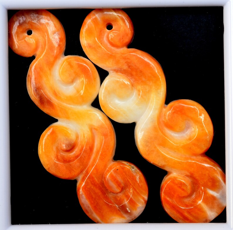 May include: Two orange and white carved jade pendants in the shape of swirling clouds. The pendants have a hole at the top for hanging.
