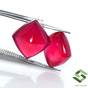 Op de afbeelding: Twee felroze vierkante edelstenen die met een pincet worden vastgehouden. De edelstenen zijn glad en glanzend.