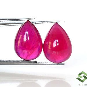 Könnte beinhalten: Zwei tropfenförmige Rubin-Edelsteine. Die Edelsteine haben eine tiefrosa-rote Farbe mit einer polierten, reflektierenden Oberfläche. Sie werden von silbernen Pinzetten gehalten und vor einem weißen Hintergrund präsentiert.