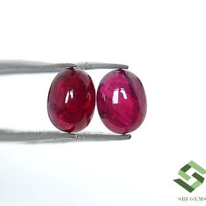 Puede incluir: Dos gemas ovaladas, pulidas, de color rojo intenso, sujetas por unas pinzas plateadas. Las gemas tienen una superficie brillante que refleja la luz. El fondo es blanco. Las gemas son probablemente rubíes, para joyería.