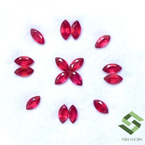 Pode incluir: Uma coleção de doze pedras preciosas vermelhas lapidadas em marquise, dispostas num padrão circular com um grupo de quatro pedras no centro. As pedras são de um vermelho vibrante com facetas visíveis. O logótipo da SBI GEMS está no canto inferior direito.