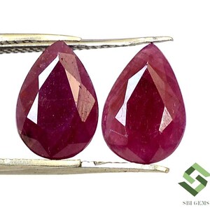 May include: Two pear-shaped ruby gemstones, each with a deep red colour and a faceted cut. The gemstones are held by a pair of tweezers.