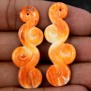 May include: Two orange and white carved shell pendants in a swirl design. The pendants are about 2 inches long and have a hole at the top for stringing.