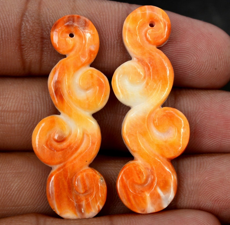 May include: Two orange and white carved shell pendants in a swirl design. The pendants are about 2 inches long and have a hole at the top for stringing.