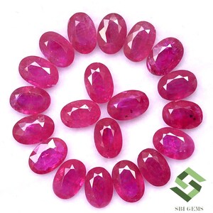 May include: A collection of 18 oval-shaped ruby gemstones, arranged in a circle on a white background. The rubies are a deep red color and have a polished finish.