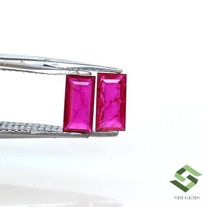 May include: Two vibrant pink rectangular ruby gemstones held by tweezers. The gemstones are cut and polished with a smooth, reflective surface.