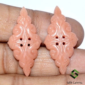 May include: Two peach-colored, carved gemstone buttons with an ornate, symmetrical design. Each button features a central four-hole pattern and a pointed top. The buttons are displayed against a neutral background.