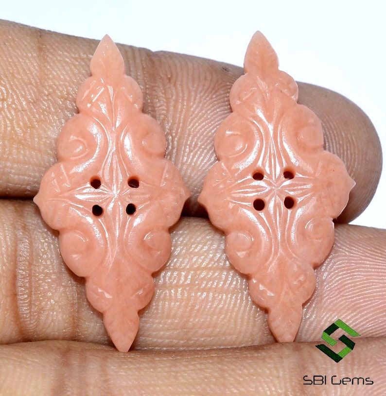 May include: Two peach-colored, carved gemstone buttons with an ornate, symmetrical design. Each button features a central four-hole pattern and a pointed top. The buttons are displayed against a neutral background.