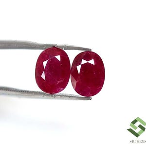 May include: Two oval-cut, deep red rubies are held by a silver tweezer against a white background. The gemstones have multiple facets, reflecting light. The image is well-lit, highlighting the rubies' colour and clarity.