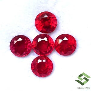 Puede incluir: Cinco gemas redondas, facetadas, de color rojo brillante dispuestas en forma de cruz. Las gemas tienen un alto brillo y están sobre un fondo blanco. Las gemas son probablemente rubíes y son adecuadas para la fabricación de joyas.