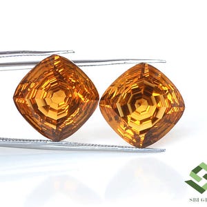 May include: Two faceted, cushion-cut gemstones with a warm, golden-orange hue. The gems are held by tweezers against a white background. The intricate cuts create a dazzling display of light and shadow, showcasing the gems' brilliance.