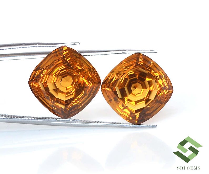 May include: Two faceted, cushion-cut gemstones with a warm, golden-orange hue. The gems are held by tweezers against a white background. The intricate cuts create a dazzling display of light and shadow, showcasing the gems' brilliance.