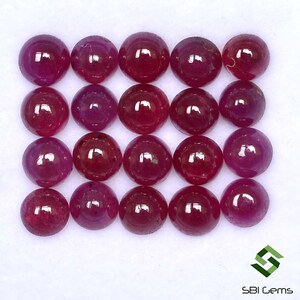 Puede incluir: Una colección de 18 cabujones redondos de rubí rojo. Los cabujones están dispuestos en una cuadrícula sobre una superficie blanca.