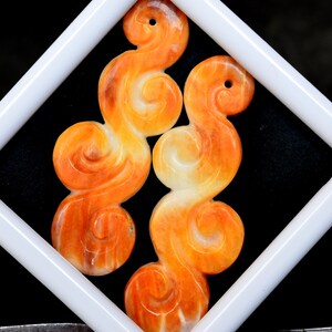 May include: Two orange and white carved jade pendants in the shape of swirling clouds. The pendants are displayed in a white square frame against a black background.