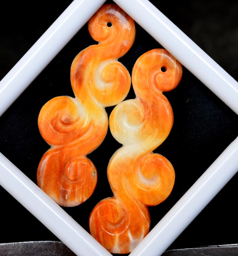 May include: Two orange and white carved jade pendants in the shape of swirling clouds. The pendants are displayed in a white square frame against a black background.