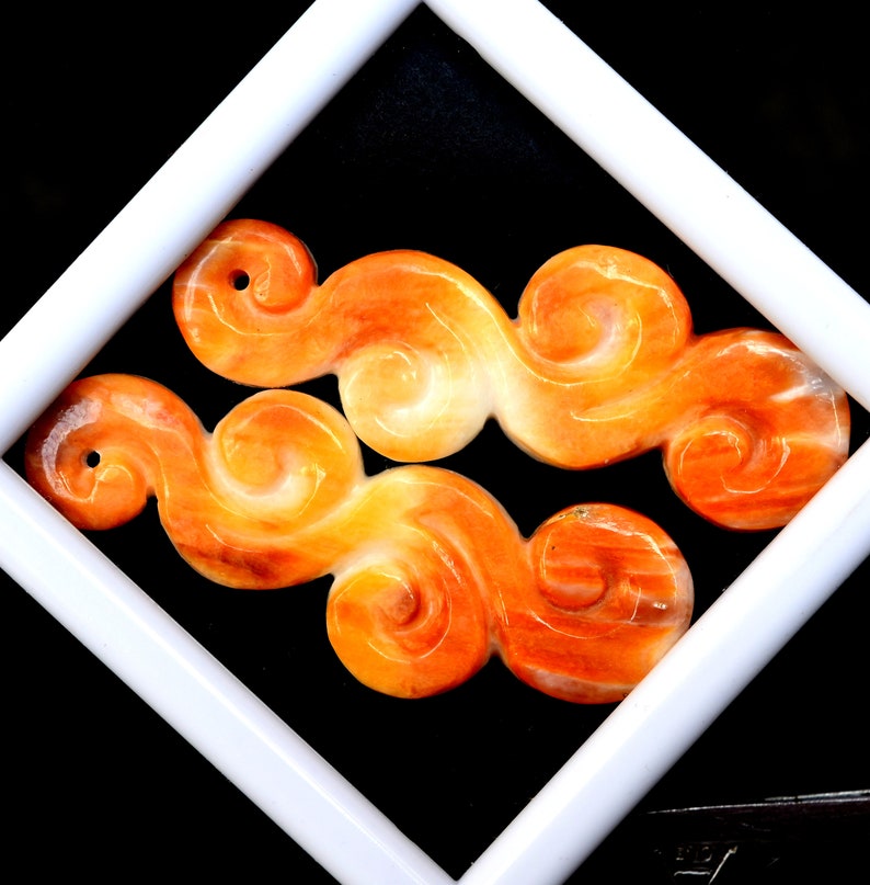 May include: Two orange and white carved jade pendants in the shape of clouds. The pendants are smooth and polished.