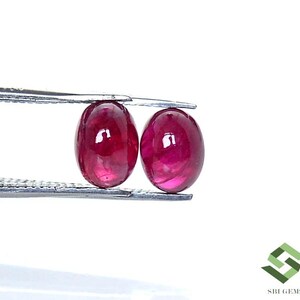 May include: Two oval-shaped, deep red gemstones are held by tweezers. The gems have a polished, glossy surface, reflecting light. The background is plain white, highlighting the vibrant colour of the rubies.