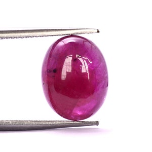 May include: A vibrant, deep pink oval-shaped gemstone with a smooth, polished surface. The gemstone is held in place by a pair of tweezers.