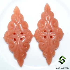 May include: Two peach-colored, carved gemstone pendants. Each pendant features an ornate, symmetrical design with floral and scroll motifs, and three small holes. The pendants are displayed on a white surface.